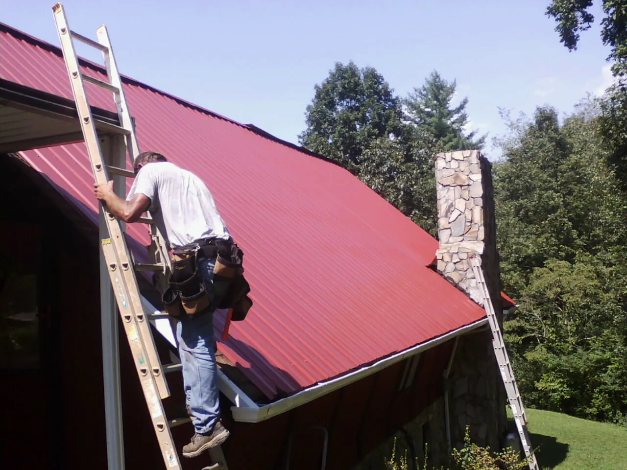 Roofer on a ladder beside a red metal roof and stone chimney — Soffit & Fascia Repair by Roofing Brothers in Madera
